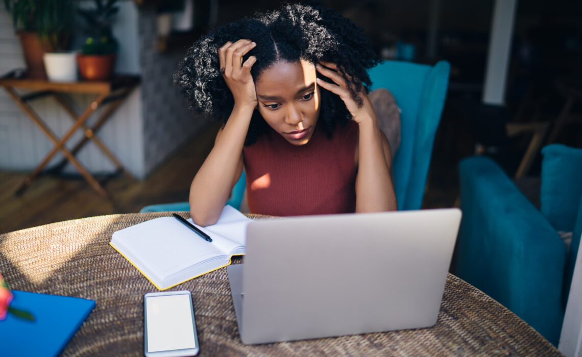Woman with head in hands looking at laptop with open notebook and smartphone on table