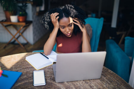 Woman with head in hands looking at laptop with open notebook and smartphone on table